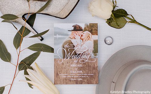 Invite to Abigail and Dalton's spring wedding surrounded by her shoe, his hat, a white rose, and eucalyptus branch. Photo by Katelyn Bradley Photography