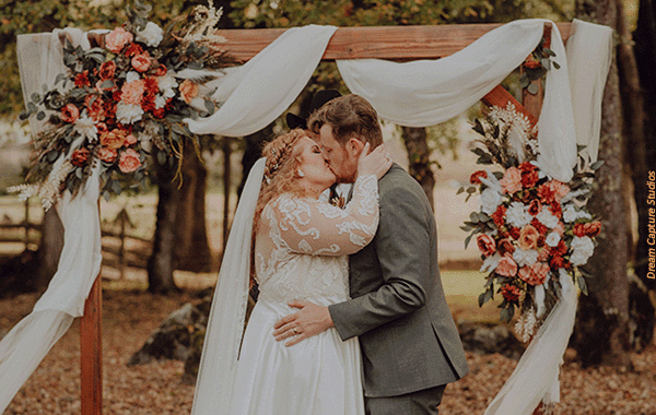 A bride and groom kissing at their ceremony with a backdrop of beautiful wedding flowers. Photo by Dream Capture Studios