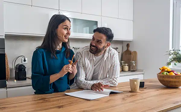 engaged-budgeting Young engaged couple standing together in their kitchen, figuring out how to plan a wedding on a budget while smiling and preparing for their upcoming celebration